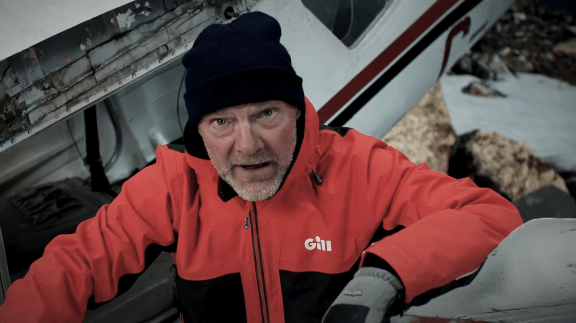 Les Strout spricht vor einem Helkopter-Wrack zu Zuschauenden.