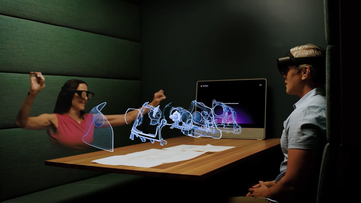 Zwei Personen mit AR-Brillen sitzen sich gegenüber und betrachten gemeinsam digitale Hologramme. Die Frau mit Brille ist selbst nur als Hologramm am Tisch und nicht als echte Person.